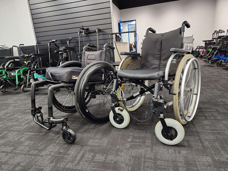 a photograph of two wheelchairs side by side. On the left is a custom carbon fibre rigid frame wheelchair, on the right is Dragancaor's broken steel folding frame generic wheelchair.