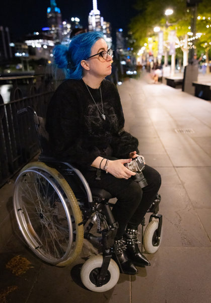 Dragancaor, a white non binary femme person with green eyes & long blue hair, sits in their wheelchair at night under pretty lights. Their hair is pulled up in a clip & they are wearing glasses, a black jumper, black jeans, & black boots with spikes.